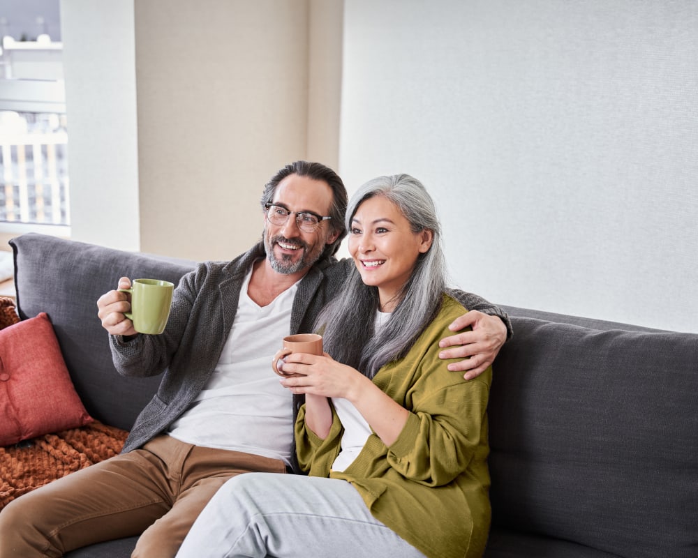 Older couple sitting on a grey couch together. The woman is holding a mug.
